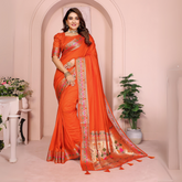 Woman wearing an orange silk saree with an intricate multicolor geometric and gold zari border, paired with a matching blouse featuring embroidered sleeves. She accessorizes with traditional jewelry including a choker necklace, bangles, and rings, and poses elegantly in a pastel archway studio with potted plants and classic decor.
