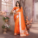 Woman wearing a vibrant orange silk saree with a contrasting gold zari border and pallu featuring traditional motifs, paired with a matching blouse. She is styled with silver jewelry and poses gracefully in a vintage-inspired floral studio with pastel blossoms, decorative vases, and a large window in the background.