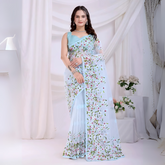 Woman wearing a pastel blue net saree with intricate multicolor floral embroidery and a matching blue sleeveless blouse, posing in a modern white studio with draped curtains and floral arrangements