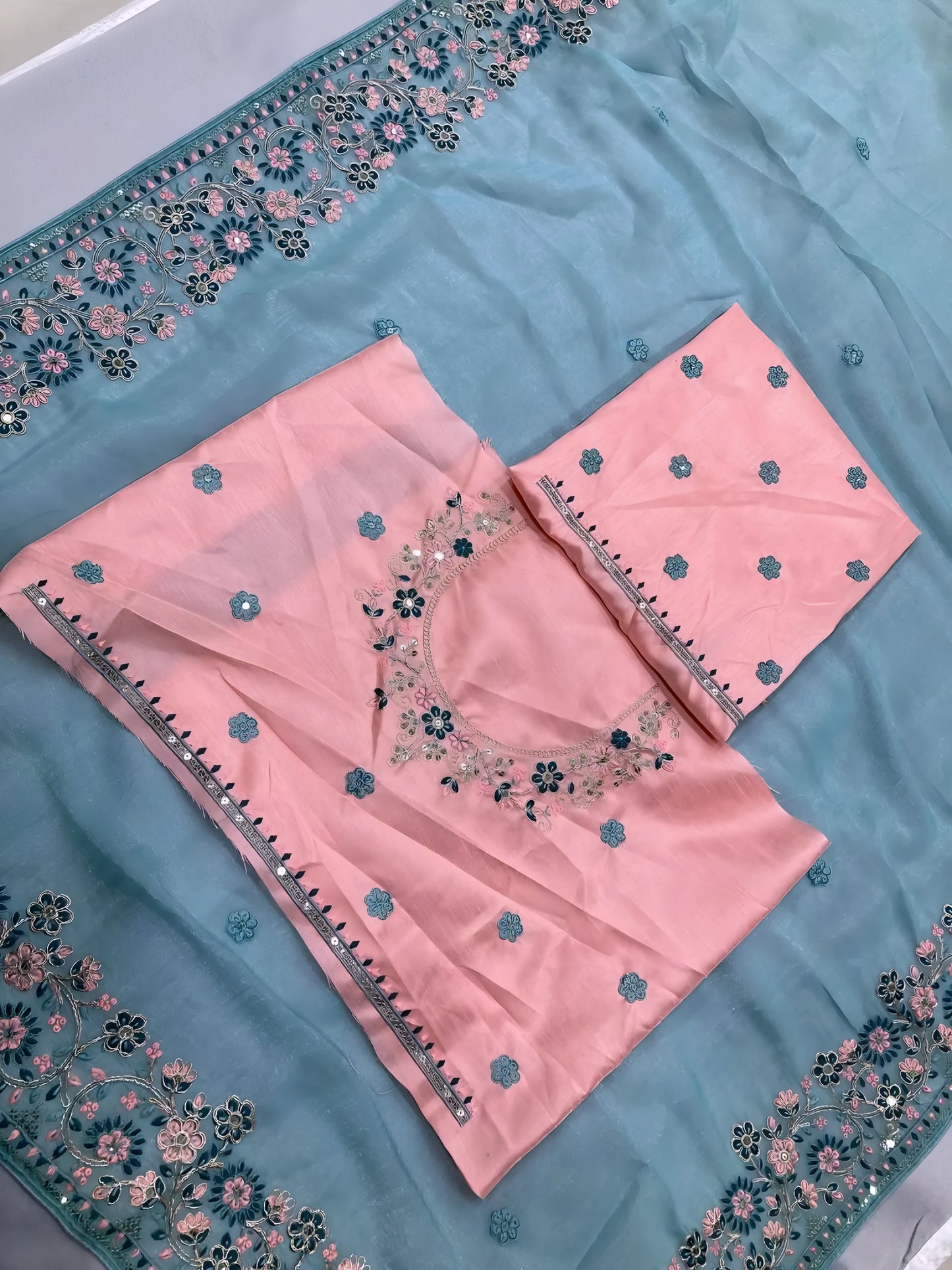 Woman wearing a pastel blue saree with intricate floral embroidery on the border, paired with a pink embroidered blouse, posing in a modern white interior with a wooden chair, decorative table, and floral arrangement.