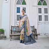 Woman wearing a pastel floral silk saree with a grey border and matching grey blouse, accessorized with traditional gold jewelry, elegantly standing in front of classic white arched doors and windows beside a vintage wooden bench, with greenery and a floral arrangement in a heritage courtyard setting.
