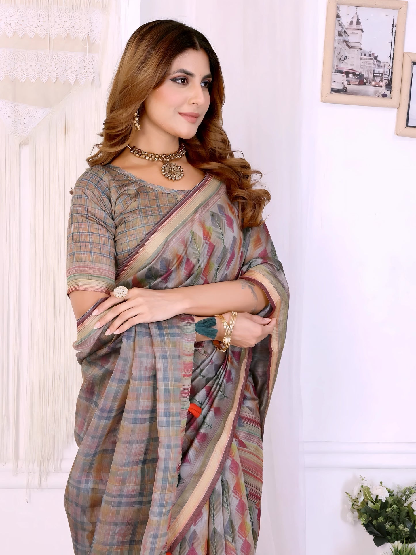 Woman wearing a pastel grey handloom saree featuring subtle multicolor geometric patterns and a beige border, styled with a matching blouse and traditional jewelry including a choker necklace and bangles. She poses elegantly in a modern room with white walls, macramé wall hanging, minimalist picture frames, and potted plants on white platforms, creating a refined and contemporary setting.