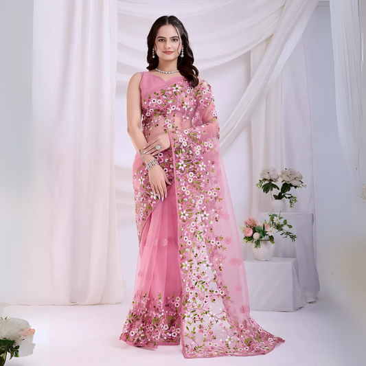 Woman wearing a pastel pink net saree with intricate white and green floral embroidery and a matching sleeveless blouse, posing in a modern white studio with draped curtains and floral arrangements.