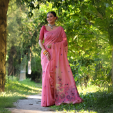Woman wearing a pastel pink saree with delicate floral embroidery and a matching pink blouse, accessorized with silver jewelry and colorful bangles, standing on a sunlit garden path surrounded by lush green trees and foliage.
