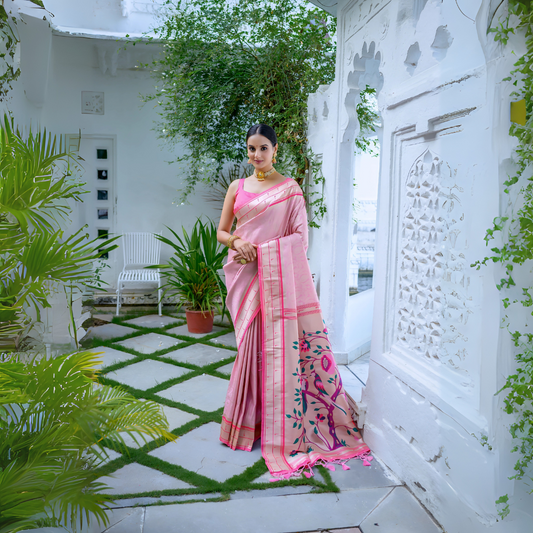Woman wearing a pastel pink silk saree with gold border and handpainted floral pallu, styled with a matching blouse and gold jewelry, posing in a white heritage courtyard surrounded by green potted plants and traditional architectural details.