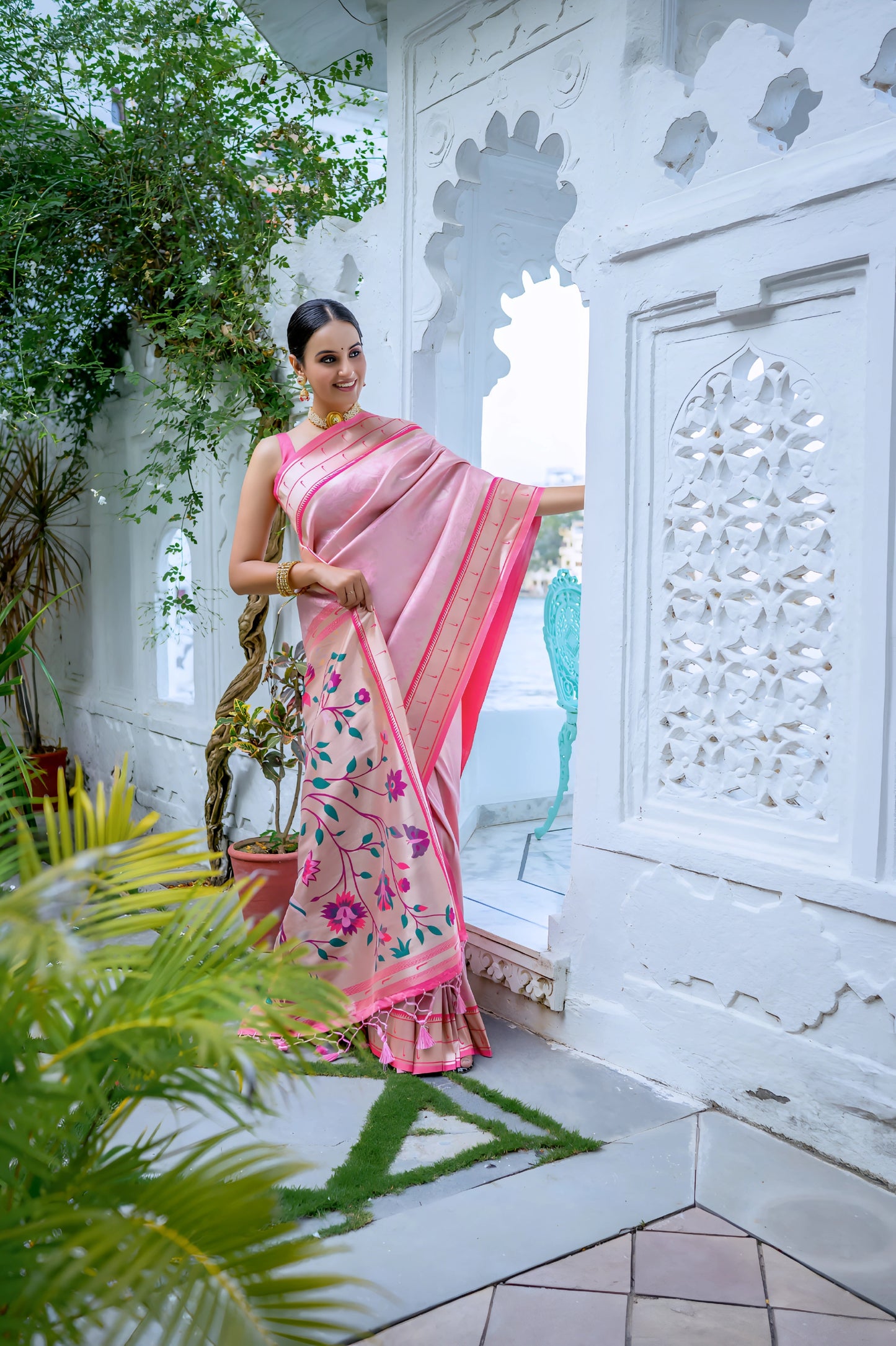 Woman wearing a pastel pink silk saree with gold border and handpainted floral pallu, styled with a matching blouse and gold jewelry, posing in a white heritage courtyard surrounded by green potted plants and traditional architectural details