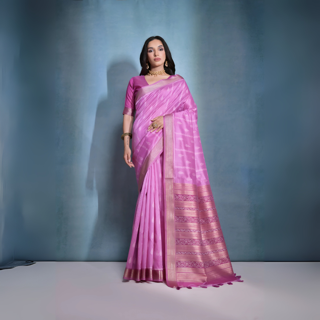 Woman wearing a pastel pink silk saree with subtle woven motifs and a gold zari border, paired with a matching blouse and traditional gold jewelry, posing against a soft blue gradient studio background.