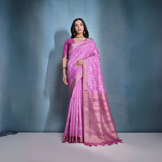 Woman wearing a pastel pink silk saree with subtle woven motifs and a gold zari border, paired with a matching blouse and traditional gold jewelry, posing against a soft blue gradient studio background.