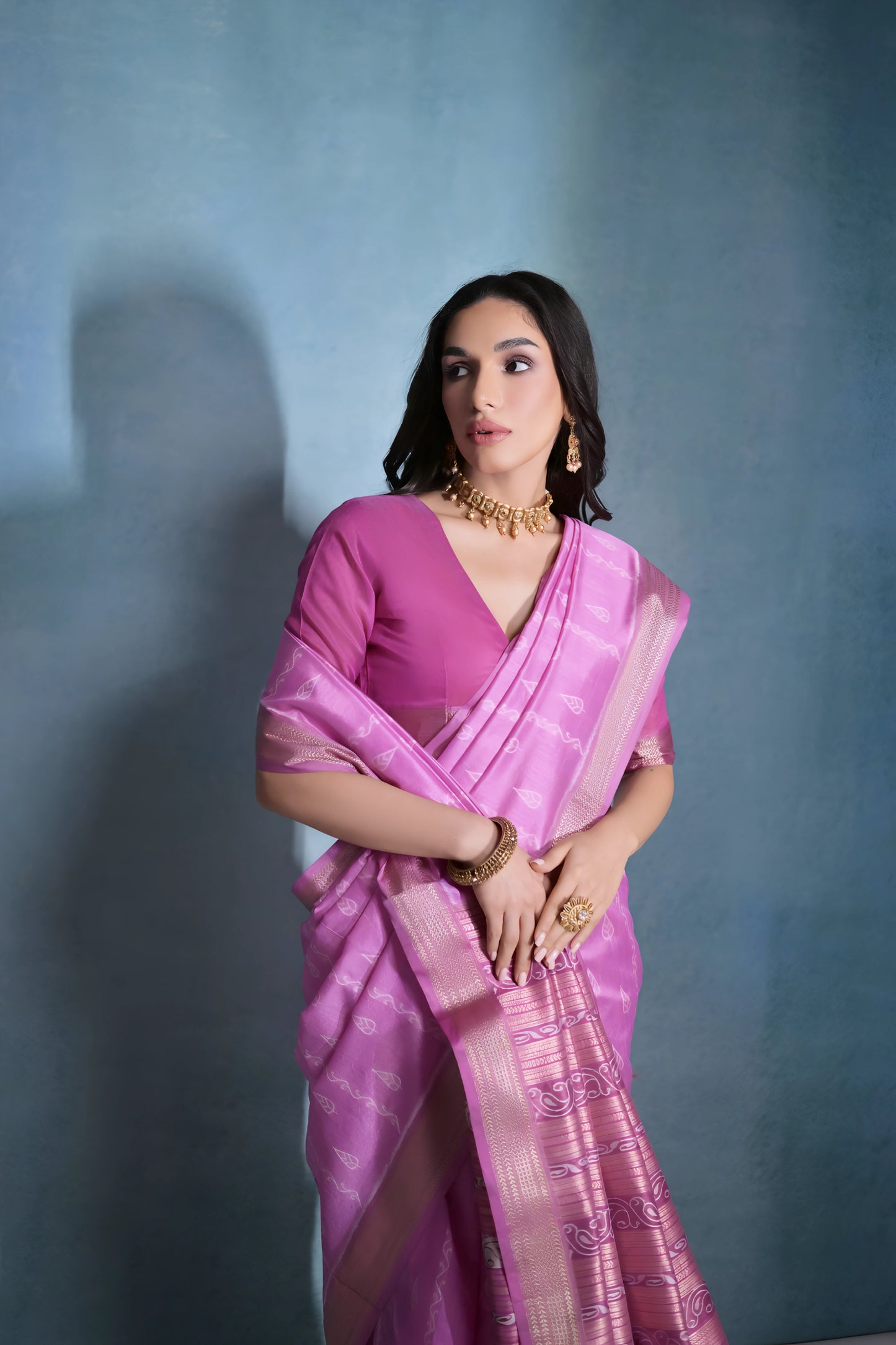 Woman wearing a pastel pink silk saree with subtle woven motifs and a gold zari border, paired with a matching blouse and traditional gold jewelry, posing against a soft blue gradient studio background.