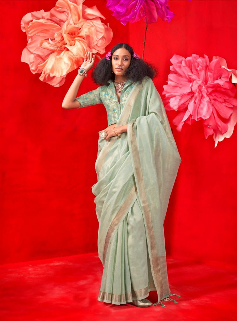 A person wearing a pastel sea green saree with intricate zari work, accessorized with bangles and a necklace, standing against a vibrant red background with large, colorful flowers.