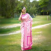 Woman wearing a pastel pink silk saree with floral prints and a decorative border, accessorized with traditional gold jewelry and bangles, standing elegantly on a grassy garden path surrounded by lush greenery and trees.
