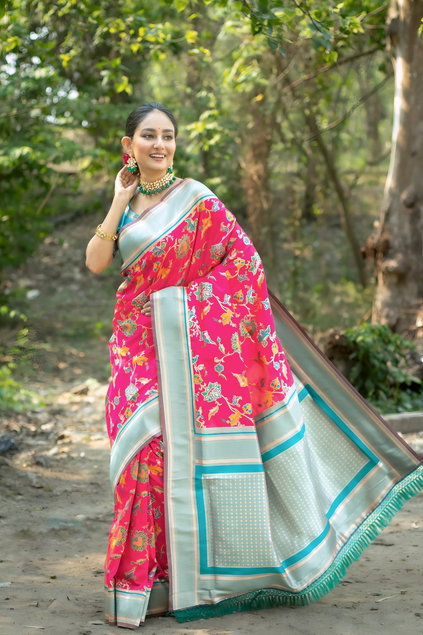 Woman wearing a vibrant pink silk saree with multicolor floral motifs and a contrasting silver border, paired with a turquoise blue blouse and traditional gold jewelry, standing gracefully on a shaded garden path with lush green trees and an open field in the background.
