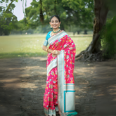 Woman wearing a vibrant pink silk saree with multicolor floral motifs and a contrasting silver border, paired with a turquoise blue blouse and traditional gold jewelry, standing gracefully on a shaded garden path with lush green trees and an open field in the background.