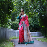 Woman wearing a vibrant pink Madhubani-printed saree with intricate traditional motifs and a contrasting green blouse, accessorized with jewelry and a long braid, standing gracefully on a garden path lined with a white fence and surrounded by lush green foliage.