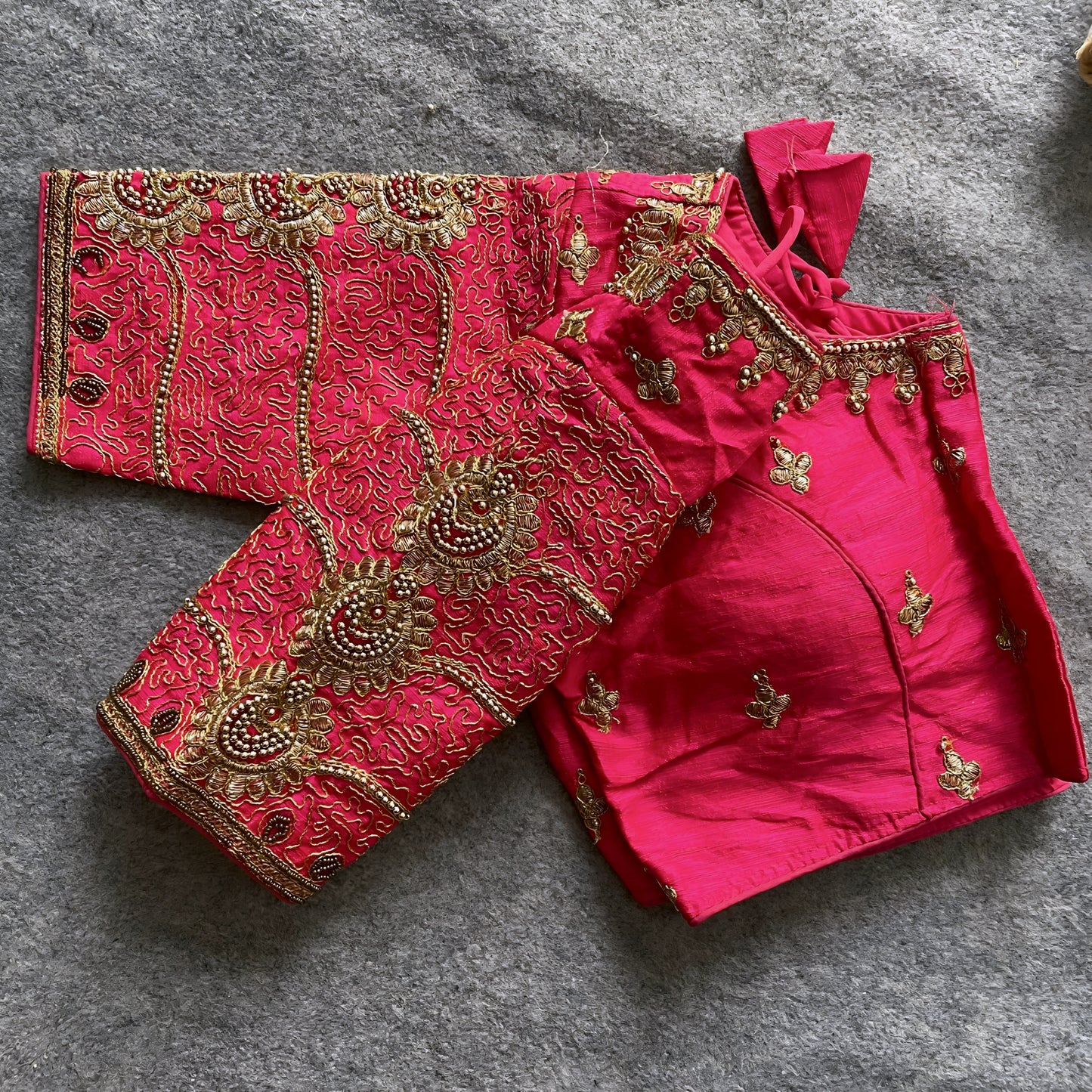 Traditional pink silk blouse featuring intricate gold embroidery and beadwork with floral and paisley motifs on the sleeves and neckline, displayed flat on a textured surface with a bunch of artificial flowers and a woven mat in the background.