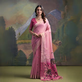 Woman wearing a pastel pink silk saree with delicate floral embroidery and a contrasting pink blouse, standing gracefully in front of a painted mountain and forest backdrop, accessorized with traditional jewelry.