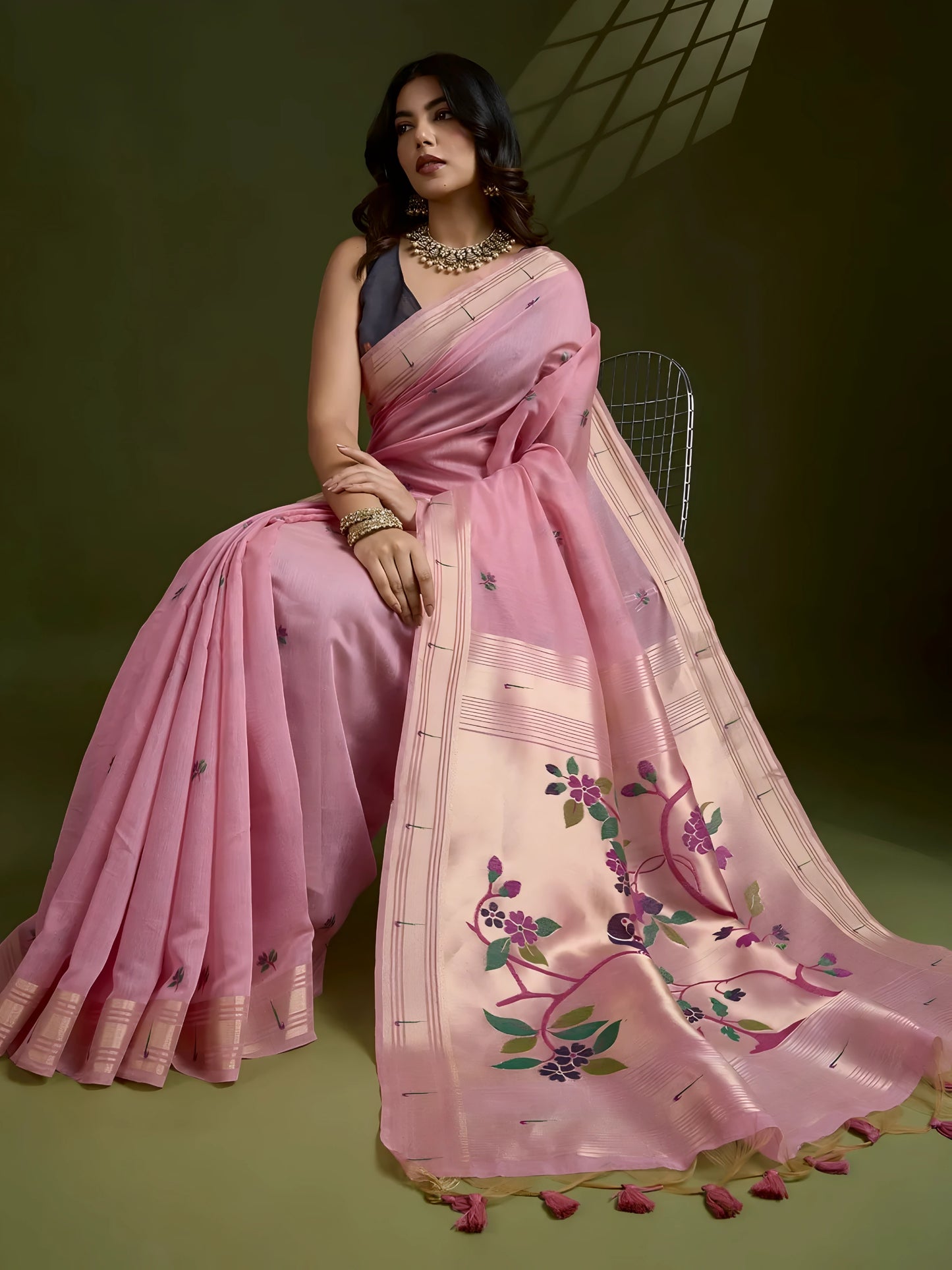 Woman wearing a pastel pink silk saree with subtle woven motifs and a contrasting pallu adorned with floral embroidery, paired with a sleeveless blouse and traditional jewelry, gracefully posing in a studio with olive green walls and patterned window light.Woman wearing a pastel pink silk saree with subtle woven motifs and a contrasting pallu adorned with floral embroidery, paired with a sleeveless blouse and traditional jewelry, gracefully posing in a studio with olive green walls and patterned window.