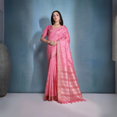 Woman wearing a pink silk saree with intricate woven patterns and gold border, paired with a matching blouse and traditional gold jewelry including a choker necklace and earrings, posing elegantly in a studio setting with a soft blue-grey gradient background.