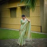 Woman in pista-colored zari work saree standing outdoors near a yellow building and palm leaves