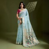 Woman wearing a powder blue silk saree with an off-white floral embroidered pallu and gold zari border, paired with a contrasting magenta blouse and traditional jewelry, elegantly posing in a studio with olive green walls and patterned window light.