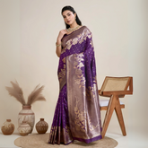 Woman wearing a rich purple Banarasi silk saree with intricate gold floral zari border and pallu, paired with a matching blouse and traditional jewelry, elegantly posing beside a wooden chair in a minimalist studio with neutral decor and ceramic vases.