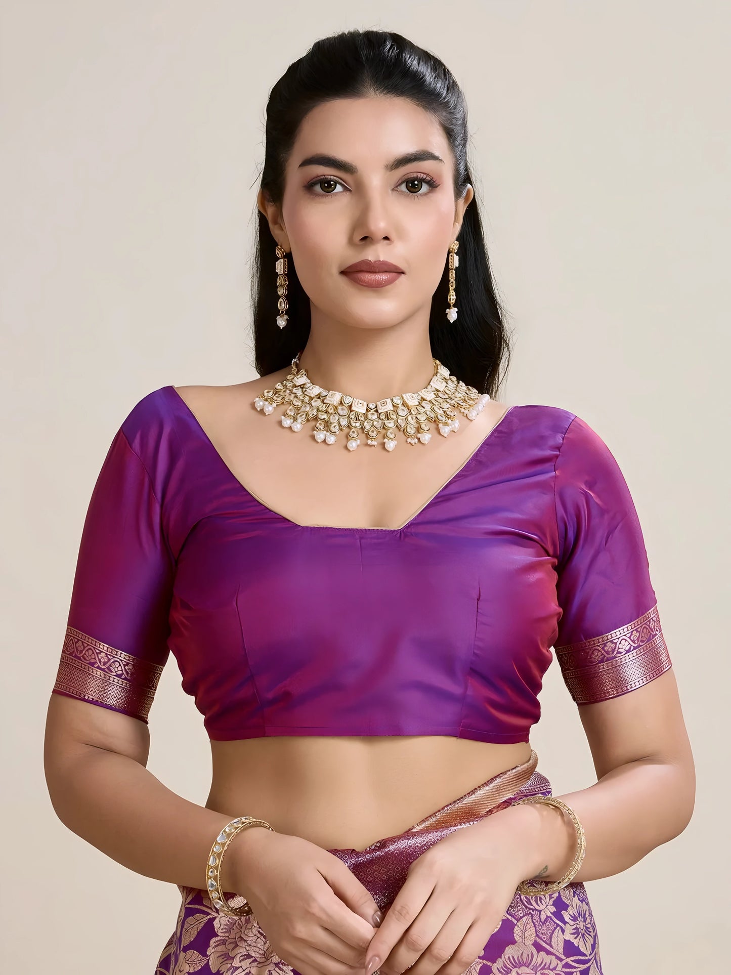Woman wearing a purple Banarasi silk saree with intricate gold floral zari border and pallu, paired with a matching blouse and traditional jewelry, elegantly posing beside a wooden chair in a minimalist studio with neutral decor and ceramic vases.