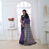 Woman wearing a purple and black Ikat printed saree with geometric patterns on the pallu, styled with a matching blouse and statement necklace, standing in a modern white interior with large arched windows and a white console table with a floral vase.