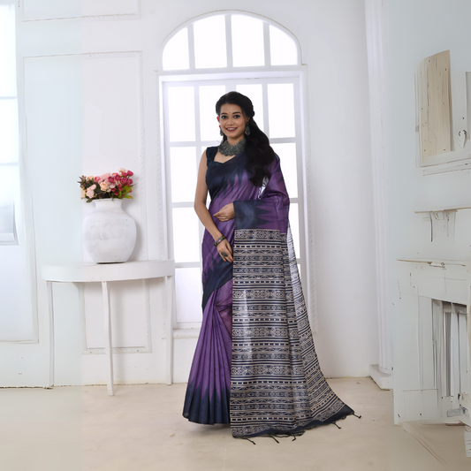 Woman wearing a purple and black Ikat printed saree with geometric patterns on the pallu, styled with a matching blouse and statement necklace, standing in a modern white interior with large arched windows and a white console table with a floral vase.