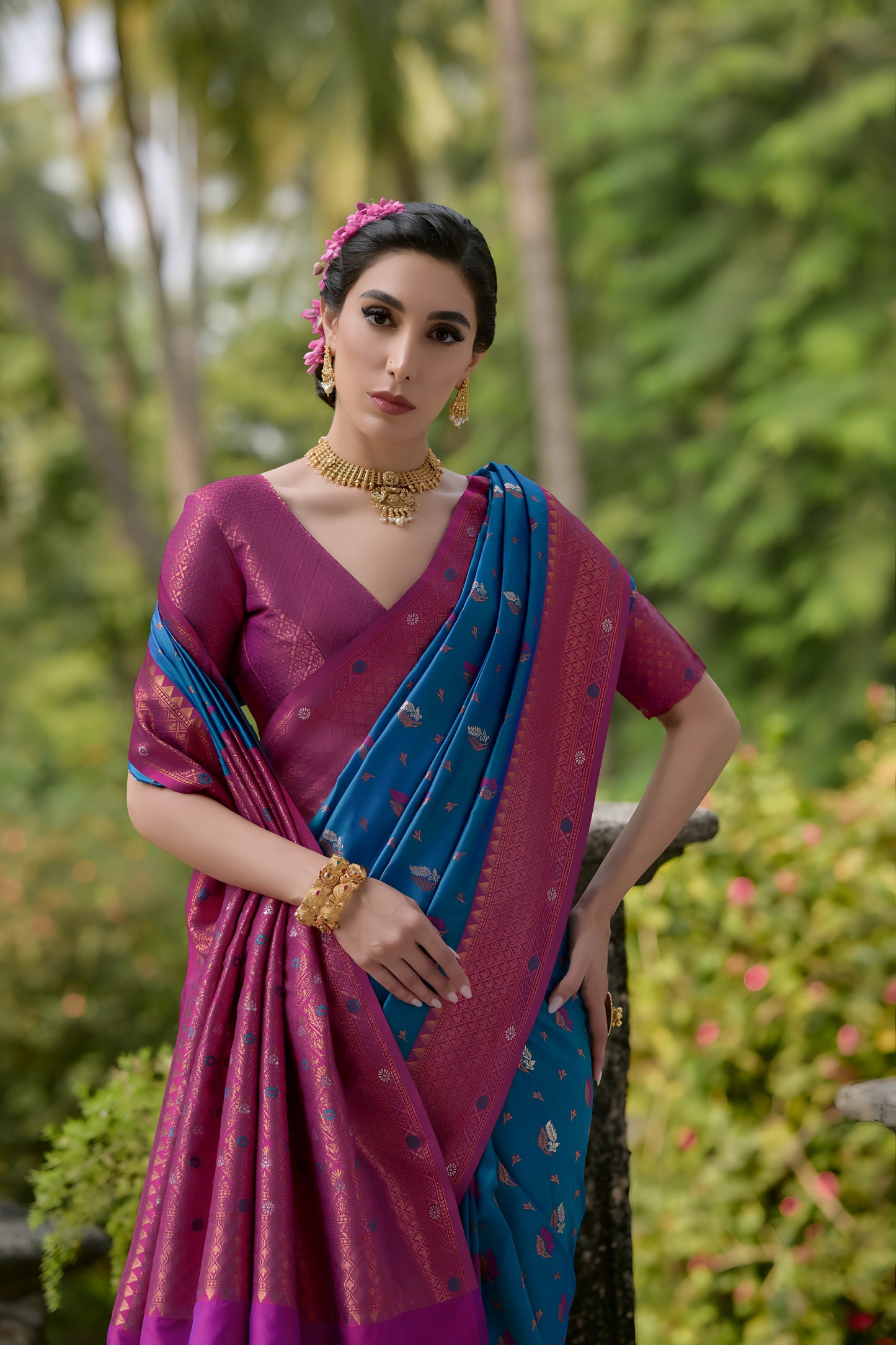Woman wearing a purple and blue silk saree with gold zari motifs and a contrasting purple border, paired with a matching blouse and traditional jewelry, standing on a vintage stone veranda surrounded by lush green trees and garden plants
