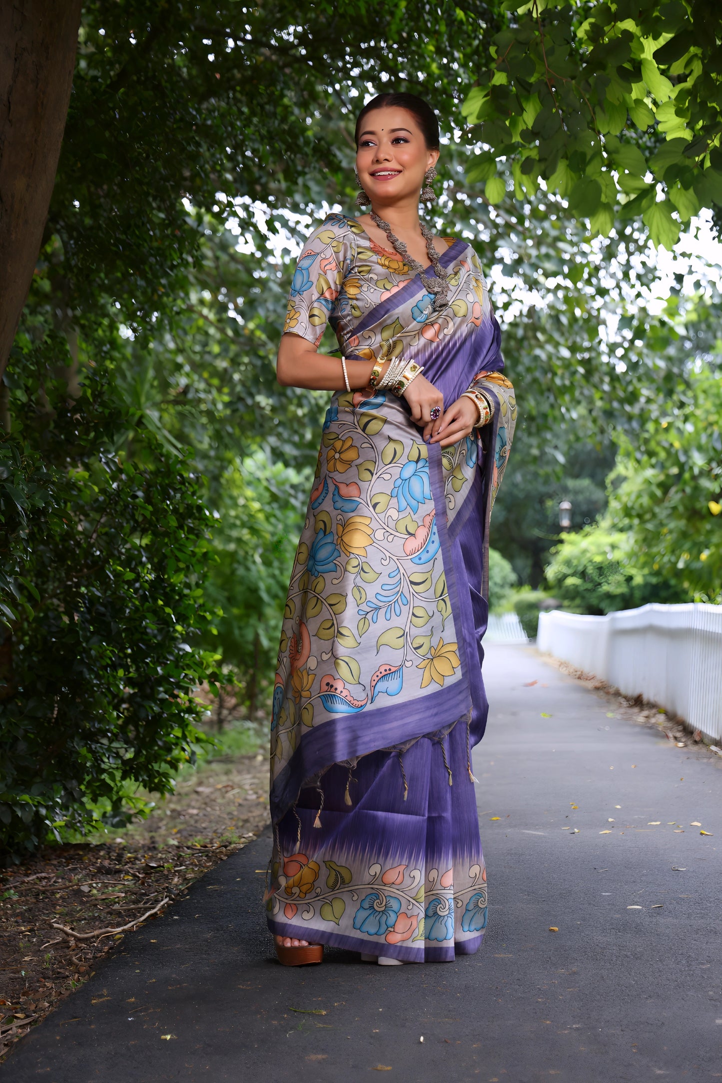 Woman wearing a purple saree with vibrant Madhubani-inspired floral print border and pallu, paired with a matching blouse and traditional jewelry, standing gracefully on a garden pathway surrounded by lush green trees and foliage.