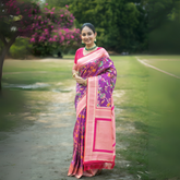 Woman wearing a vibrant purple silk saree with pink borders and multicolor floral motifs, paired with a pink blouse and traditional jewelry, standing gracefully on a garden path surrounded by lush greenery and blooming trees.