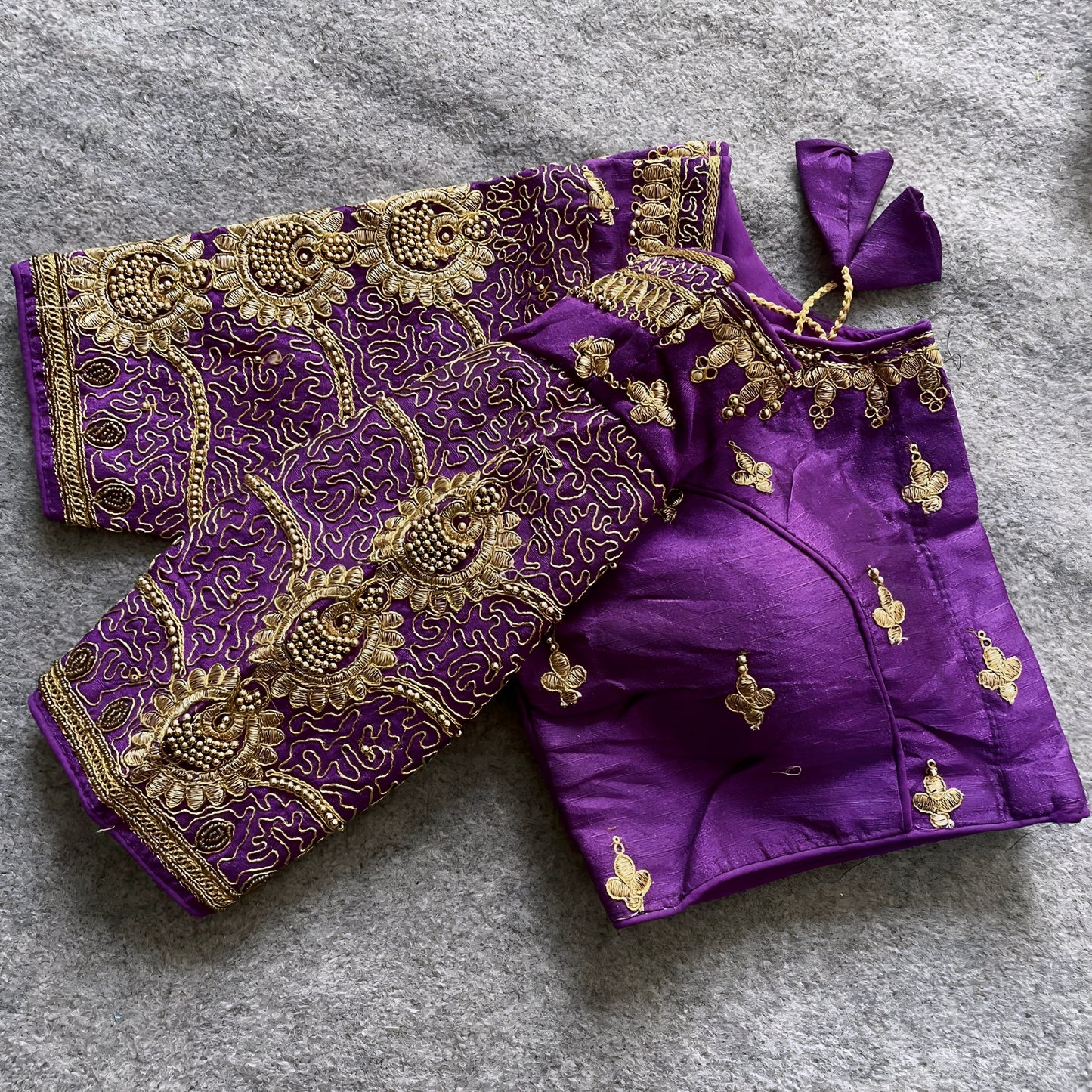 Traditional purple silk blouse featuring intricate gold embroidery and beadwork with floral and paisley motifs on the sleeves and neckline, displayed flat on a textured surface with a bunch of artificial flowers and a woven mat in the background.