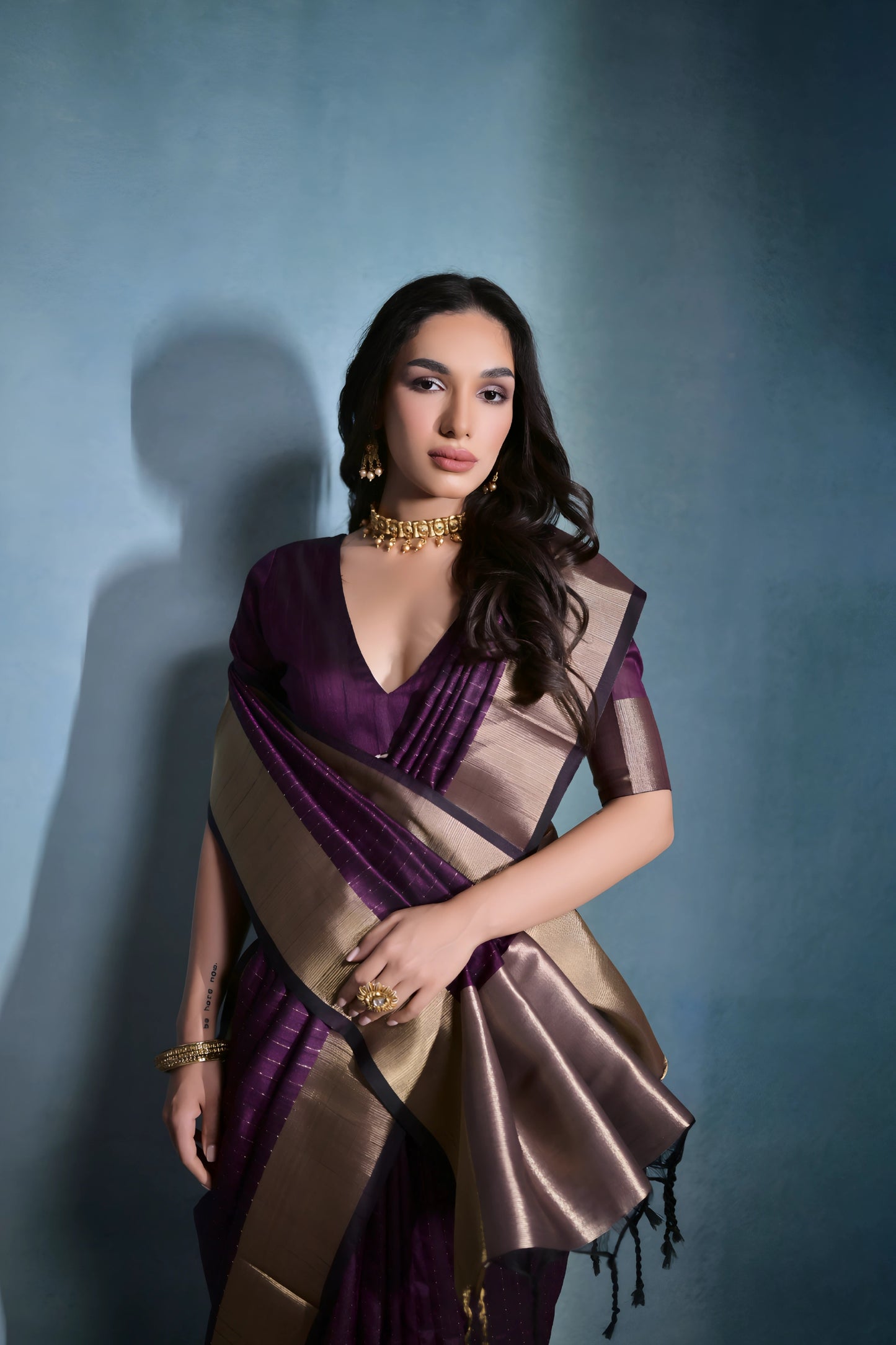 Woman wearing a purple silk saree with a gold border and traditional jewelry, standing against a blue background.