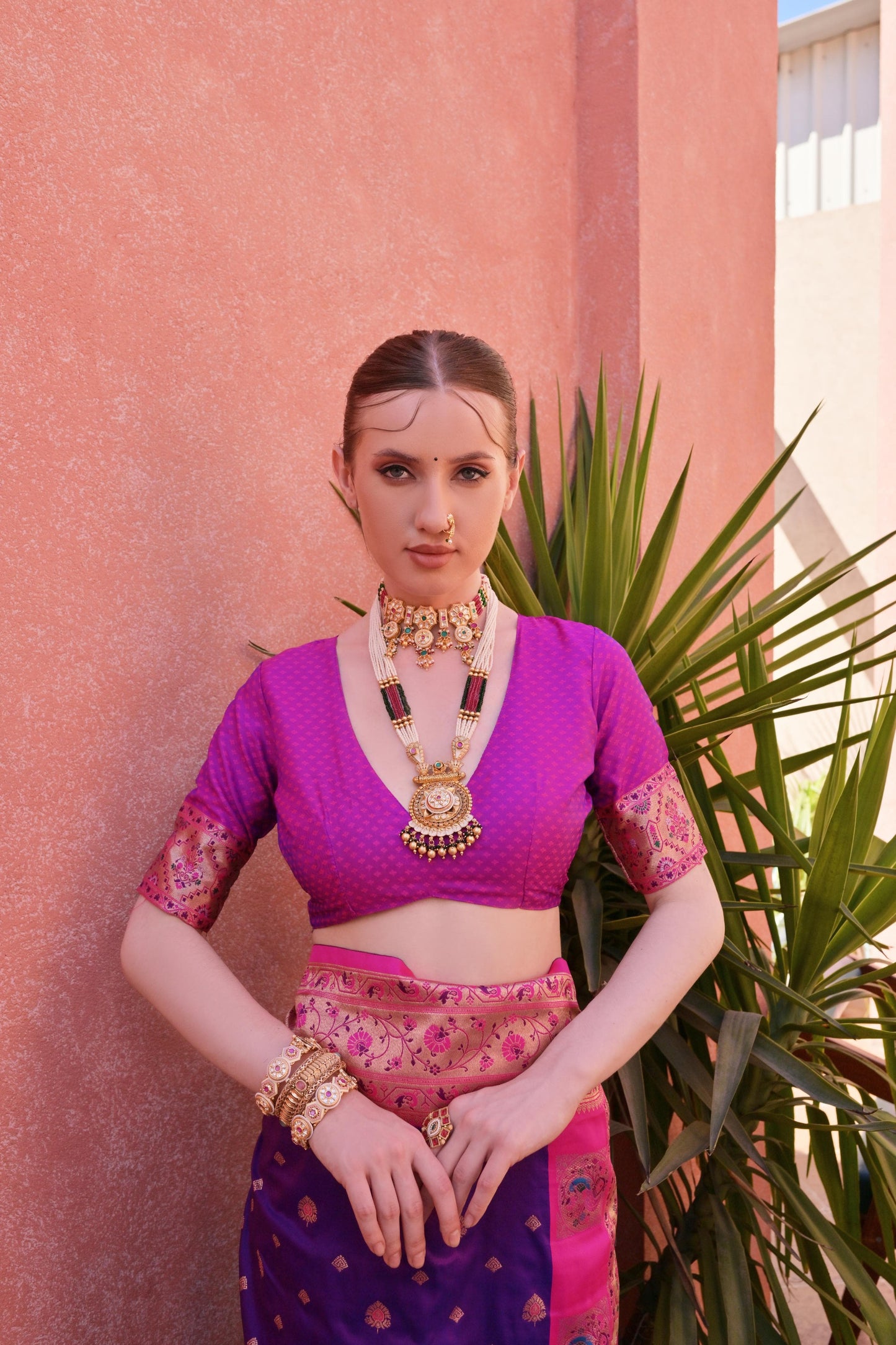 Woman wearing a purple silk saree with pink and gold border, adorned with traditional gold jewelry, standing by a terracotta wall and green plantv.