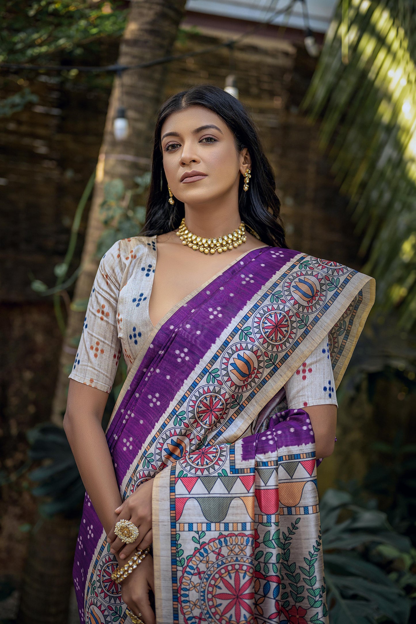 Woman wearing a purple saree with intricate white tribal-inspired prints and a multicolor pallu featuring circular and geometric patterns, paired with a short-sleeve embroidered blouse and traditional jewelry, standing gracefully in a lush outdoor garden surrounded by tropical plants.