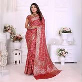 Woman draped in a vibrant red Banarasi bandhani saree featuring intricate white tie-dye patterns and an ornate zari border, paired with a matching short-sleeve blouse and traditional jewelry. She stands gracefully in a classic white room adorned with paneled walls and elegant white vases filled with pastel and rose-toned artificial flower arrangements.