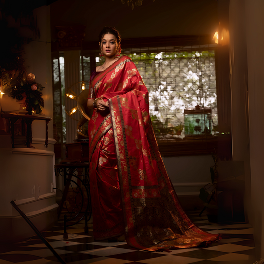 Woman wearing a red Banarasi silk saree with intricate gold zari border and floral motifs, accessorized with traditional jewelry, standing in a vintage-inspired room with checkered flooring, a gramophone, floral decor, and bamboo blinds filtering natural light.