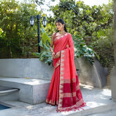 Woman wearing a red handloom silk saree with gold zari border and geometric patterns, paired with a sleeveless blouse and traditional jewelry, standing outdoors on a concrete platform surrounded by lush green plants and garden lighting.
