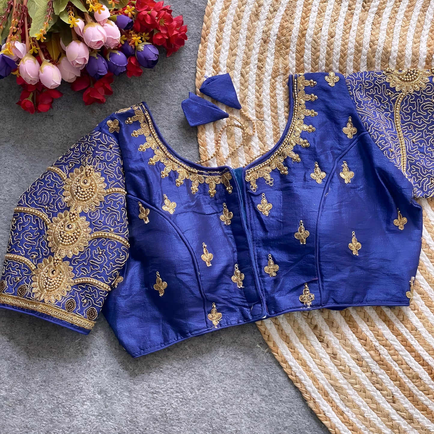 Traditional royal blue silk blouse featuring intricate gold embroidery and beadwork with floral and paisley motifs on the sleeves and neckline, displayed flat on a textured surface with a bunch of artificial flowers and a woven mat in the background.