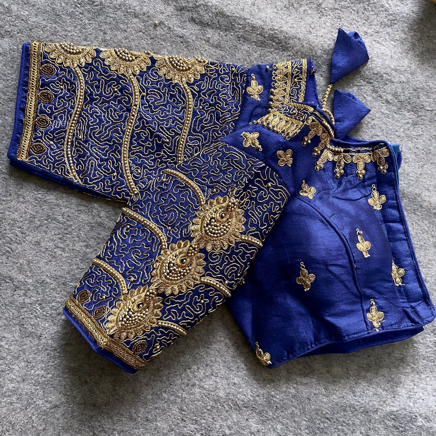 Traditional royal blue silk blouse featuring intricate gold embroidery and beadwork with floral and paisley motifs on the sleeves and neckline, displayed flat on a textured surface with a bunch of artificial flowers and a woven mat in the background.