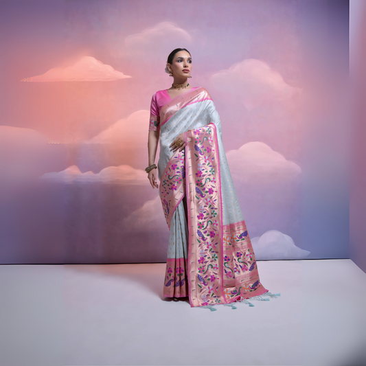 Woman wearing a silver Banarasi silk saree with an intricate pink floral zari border and pallu, paired with a pink blouse and traditional jewelry, posing in a pastel-themed studio with dreamy cloud backgrounds