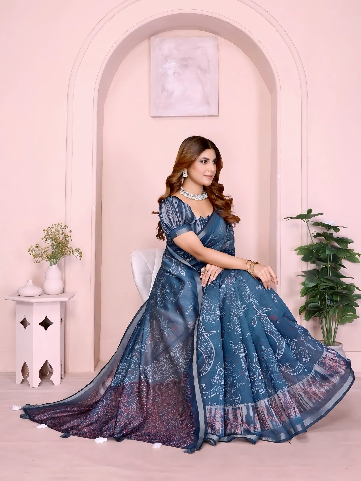 Woman wearing a steel blue printed saree with intricate abstract and paisley designs, paired with a matching short-sleeve blouse and accessorized with a silver choker necklace, bangles, and rings. She poses gracefully in a modern studio featuring a pastel arched alcove, a white side table with a ceramic vase of green foliage, and a potted plant against a soft blush background.