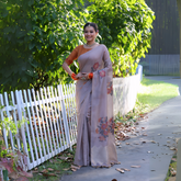 Woman wearing a taupe silk saree with subtle floral prints and an orange blouse, accessorized with pearl jewelry and orange bangles, standing beside a white picket fence in a sunlit garden with lush green foliage in the background.