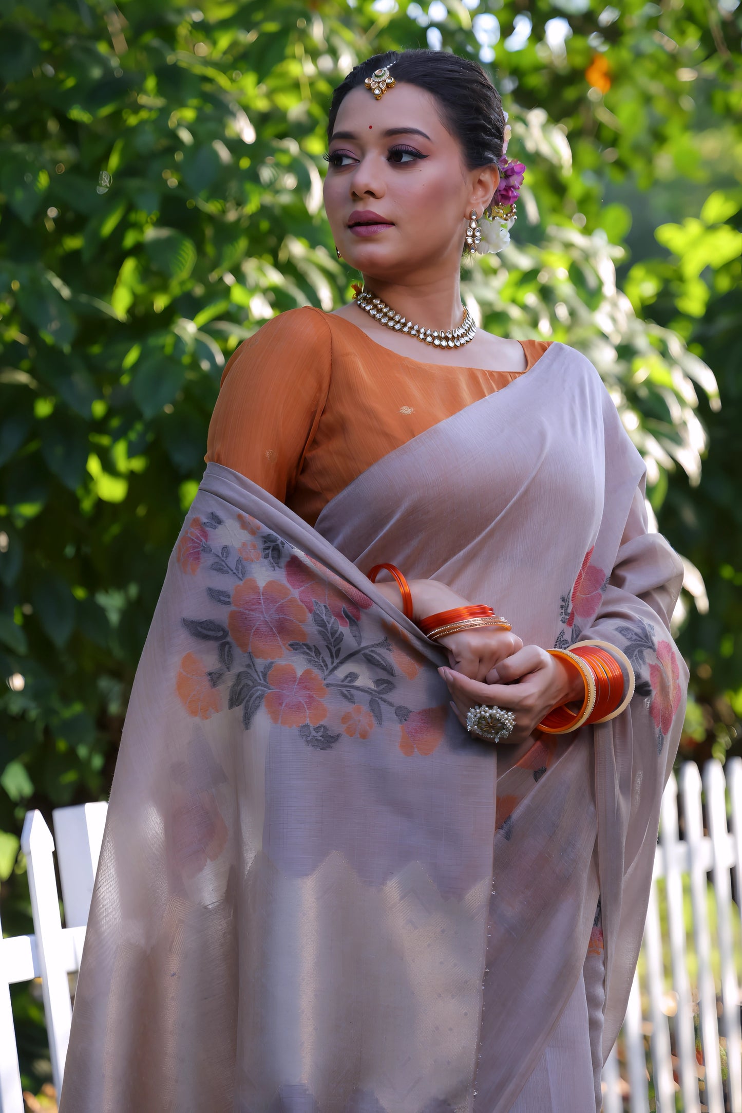 Woman wearing a taupe silk saree with subtle floral prints and an orange blouse, accessorized with pearl jewelry and orange bangles, standing beside a white picket fence in a sunlit garden with lush green foliage in the background.