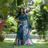 Woman wearing a teal saree with large handpainted lotus and leaf floral motifs, accessorized with traditional gold jewelry, standing on a lush green lawn surrounded by tropical plants and a white picket fence in the background