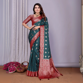 Woman draped in an elegant teal Banarasi silk saree adorned with intricate silver woven motifs and a rich red zari border, paired with a matching red blouse and classic gold jewelry. She poses gracefully in a contemporary studio decorated with woven baskets filled with rose petals, sheer white curtains, and a minimalist neutral backdrop.