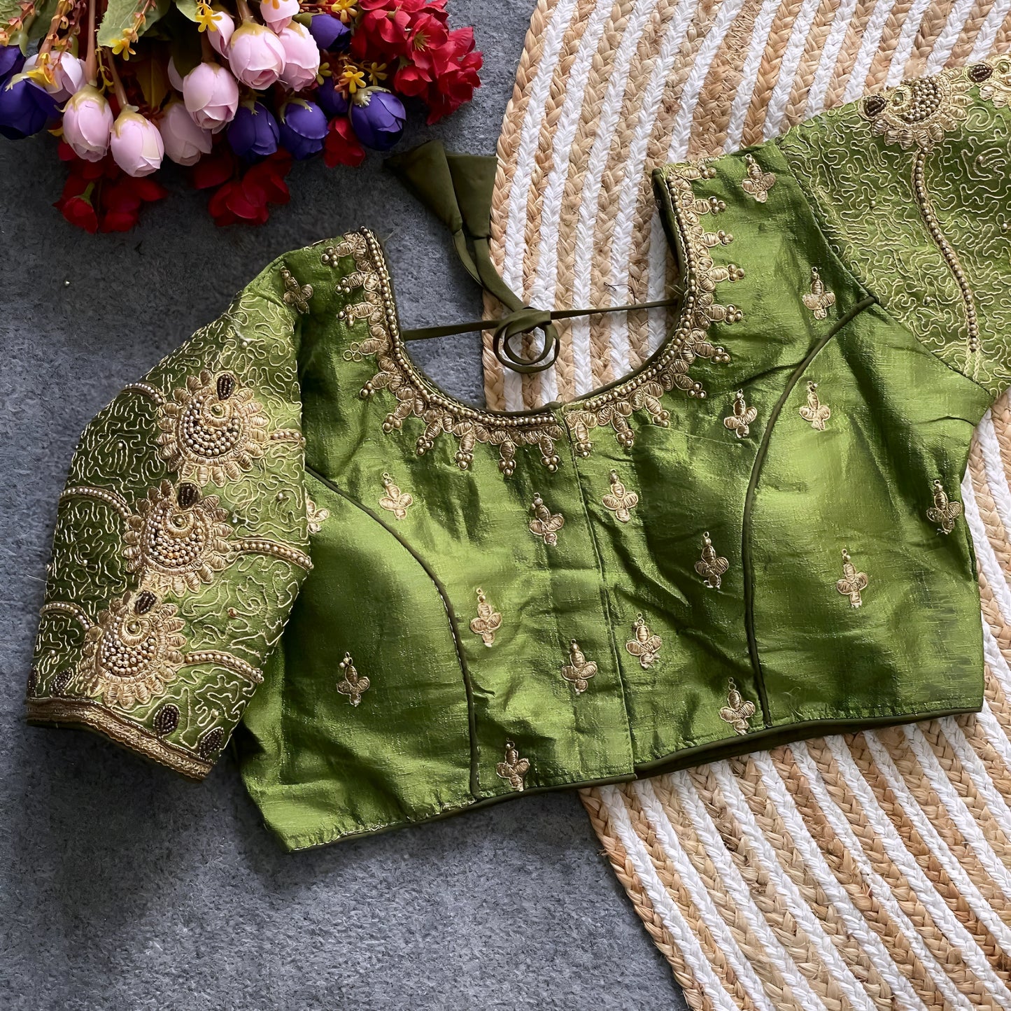Traditional teal silk blouse featuring intricate gold embroidery and beadwork with floral and paisley motifs on the sleeves and neckline, displayed flat on a textured surface with a bunch of artificial flowers and a woven mat in the background.