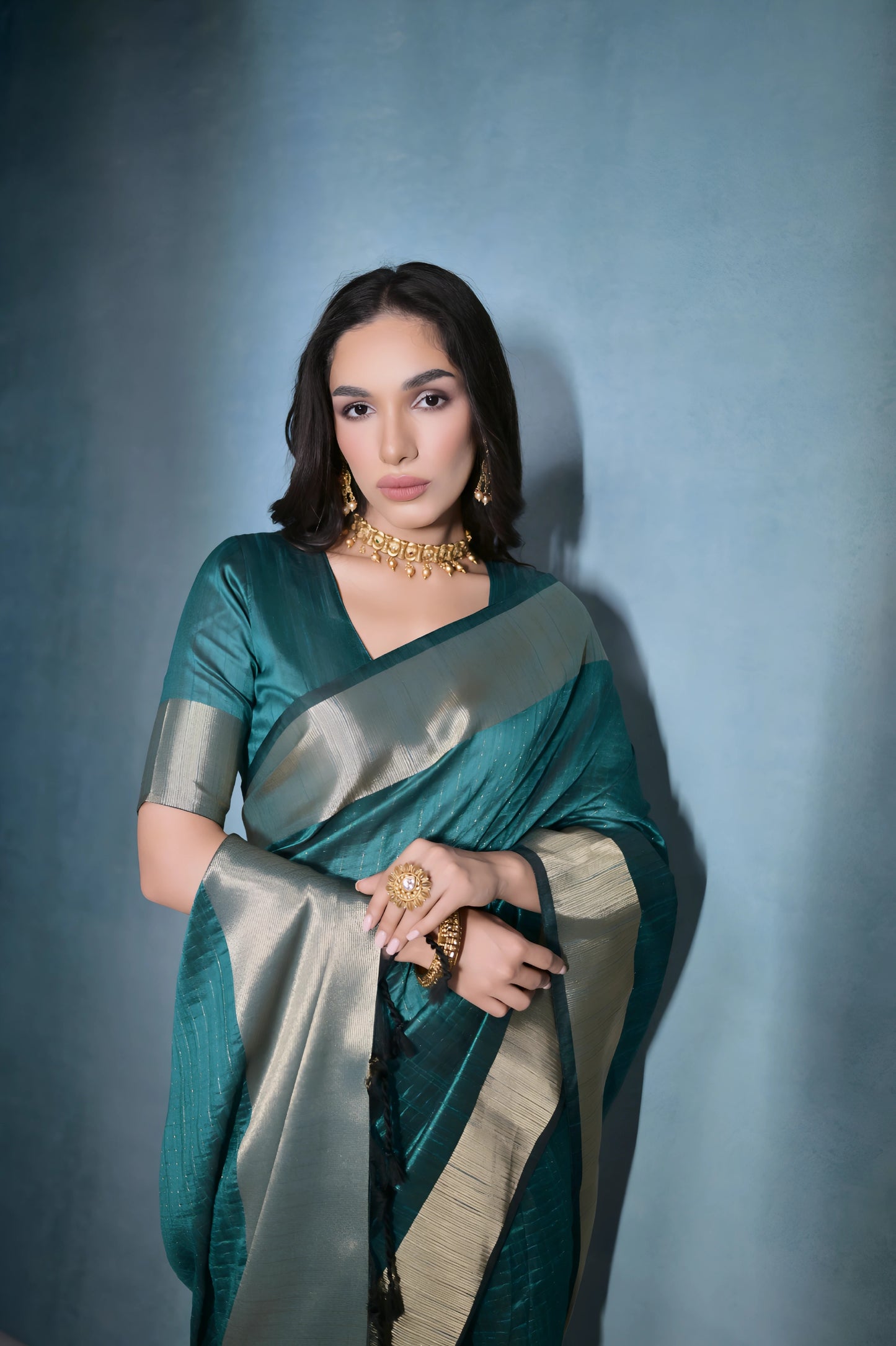 Woman wearing a teal silk saree with a gold border and traditional jewelry, standing against a blue background.
