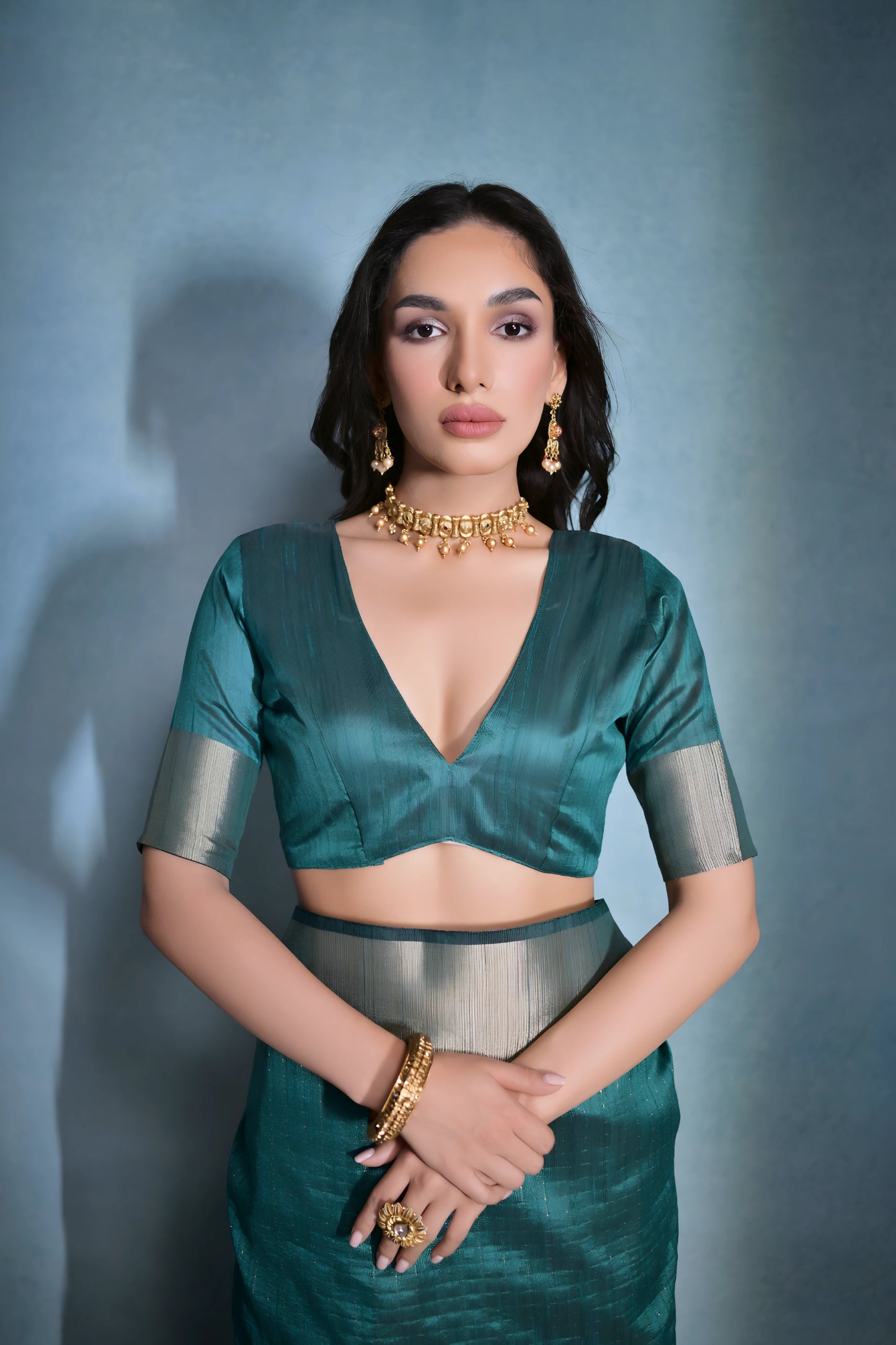 Woman wearing a teal silk saree with a gold border and traditional jewelry, standing against a blue background