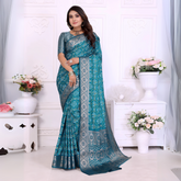 Woman wearing a teal blue silk saree with intricate silver zari border and traditional floral motifs, paired with a matching blouse and statement jewelry. She poses gracefully in a classic white room decorated with paneled walls and white vases filled with pastel floral arrangements.